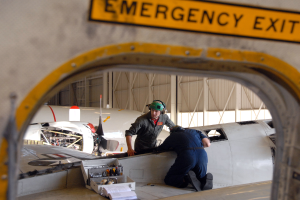 Zwei M√§nner arbeiten an einem Flugzeug in einer Halle, einer sitzt auf dem Flugzeug und tr√§gt einen Helm, der andere steht neben ihm, mit Werkzeugen und Papieren auf einem Tisch und einer Notausgangstafel an der Wand dahinter.