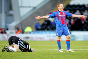 Ein Stadion mit einer Person, die steht, und einer anderen, die auf dem Rasen kniet, sowie einer Gruppe von Menschen im Hintergrund und Tafeln mit Text dahinter.