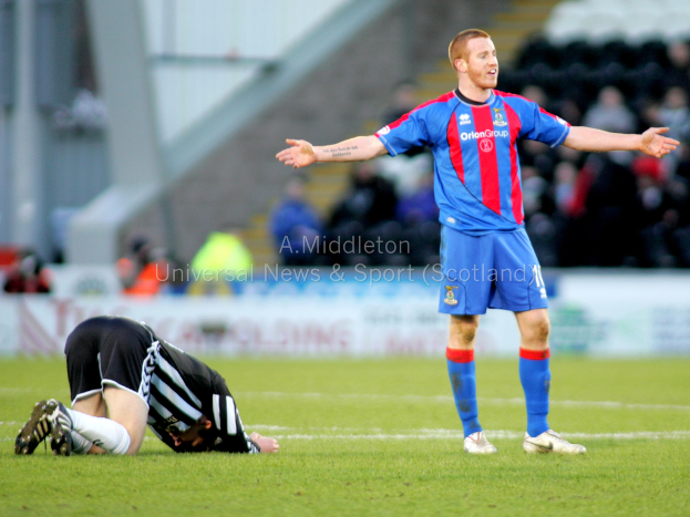Ein Stadion mit einer Person, die steht, und einer anderen, die auf dem Rasen kniet, sowie einer Gruppe von Menschen im Hintergrund und Tafeln mit Text dahinter.