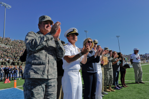Eine Gruppe von Menschen auf einem Fußballfeld klatschen, mit Zuschauern auf den Rängen, Laternen und einem klaren blauen Himmel im Hintergrund.
