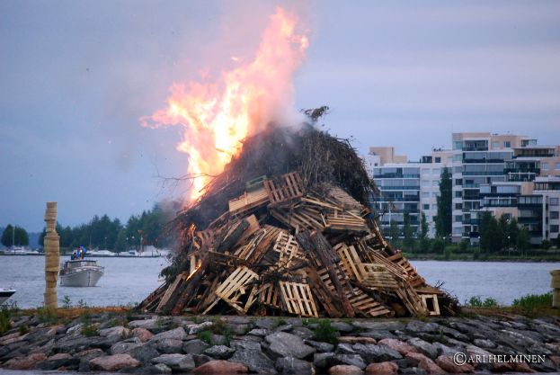 Holzobjekte brennen mit Feuer, mit Steinen unten und Wasser in der Mitte, auf dem ein Schiff schwimmt, umgeben von Gebäuden, Bäumen und einem Himmel im Hintergrund.