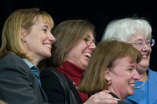 Mehrere ältere Frauen stehen in der foreground, lächeln und schauen nach rechts, mit einem schwarzen Hintergrund dahinter.