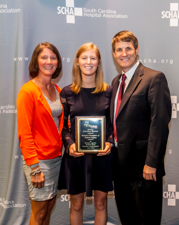 Drei Personen, zwei Frauen und ein Mann, stehen zusammen. Der Mann trägt einen schwarzen Anzug, eine Frau trägt ein orange-schwarzes Kleid und die andere Frau trägt ein schwarzes Kleid und hält einen Preis in den Händen. Sie stehen vor einem grauen Hintergrund.