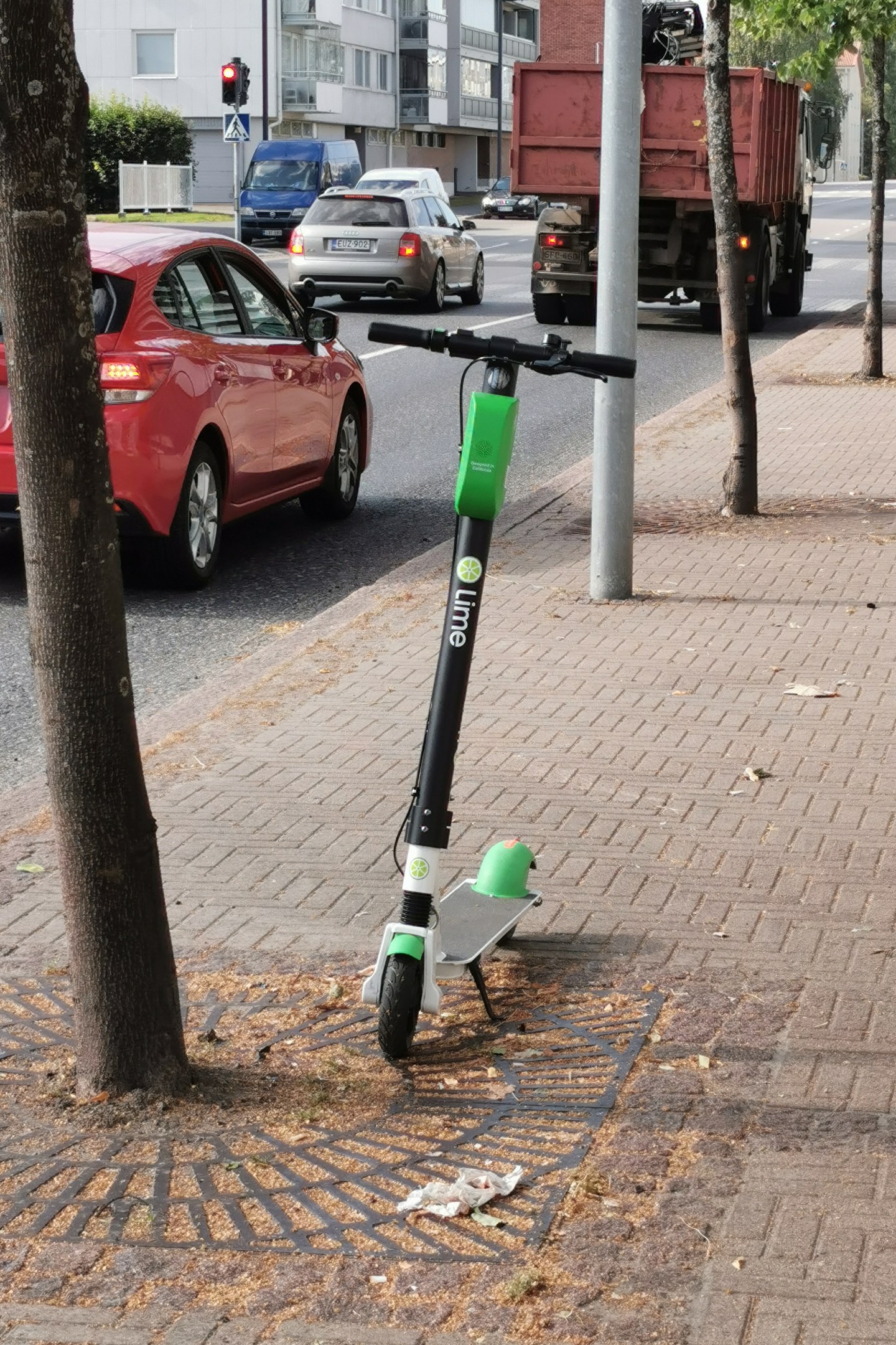 Elektroroller auf der Straße neben einem Baum geparkt, mit Fahrzeugen und Gebäuden im Hintergrund.