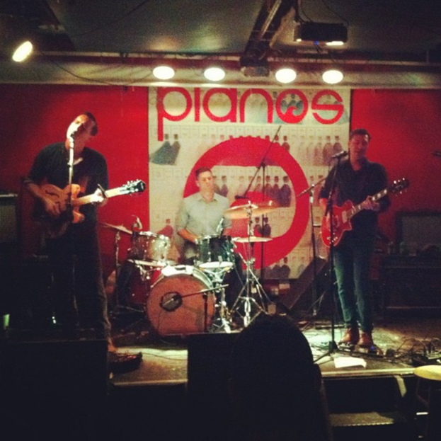 Drei Männer auf der Bühne bei einem Konzert: einer spielt am Schlagzeug in der Mitte, und zwei spielen Gitarren und singen auf beiden Seiten, mit einem Banner an der Wand und Lampen an der Decke darüber.