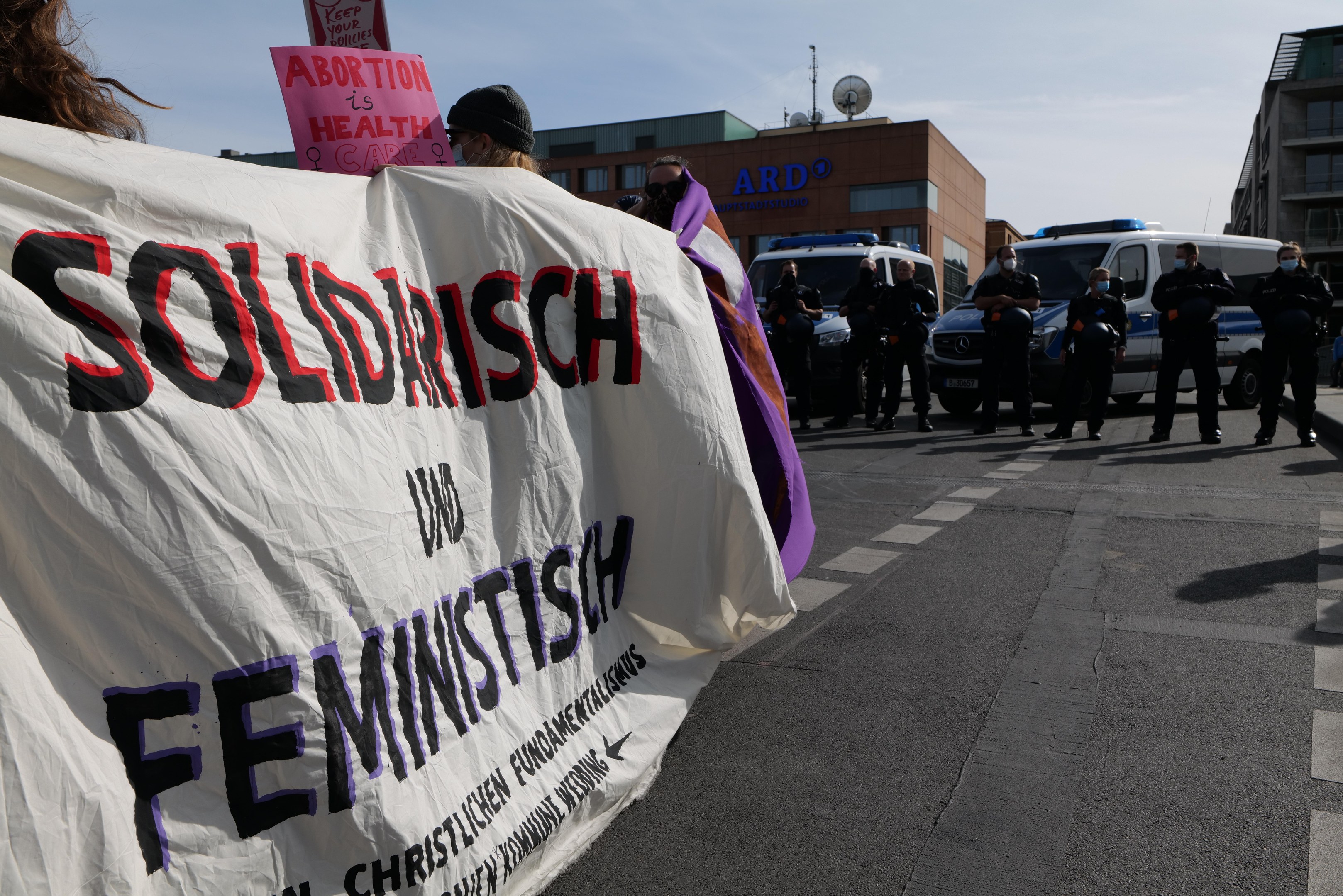 Eine Gruppe von Menschen marschiert die Straße entlang, hält ein Banner mit der Aufschrift "Solidarität und Feminismus", mit parkenden Fahrzeugen, Gebäuden, einer Schüsselantenne und einem klaren blauen Himmel im Hintergrund.