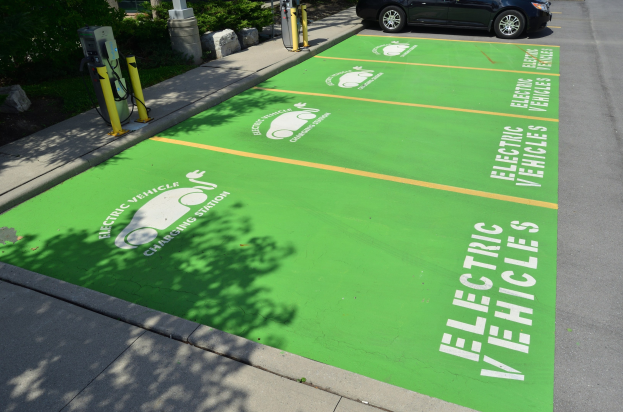 Elektroauto auf einer Ladestation mit einer grünen Bodenmatte mit Text und Bildern in einer Parkanlage umgeben von Bäumen, Pflanzen, Pfählen und anderen Gegenständen.