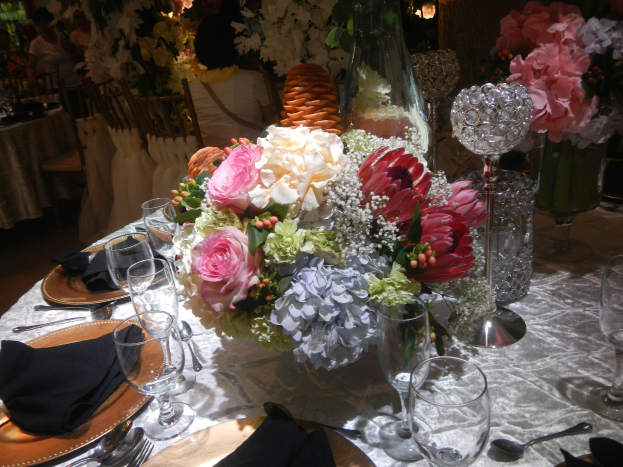 Ein formeller Esstisch mit Tellern, Besteck, Gläsern, Servietten und Blumenvasen, mit einem floralen Zentrum und im Hintergrund sitzende Gäste.