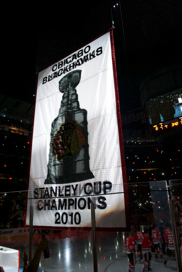 Eine nächtliche Stadionkulisse mit einem großen Banner in der Mitte, Zuschauern im Hintergrund, einem Glas auf dem Boden und ein paar Spielern auf dem Feld, beleuchtet von Stadionlampen.