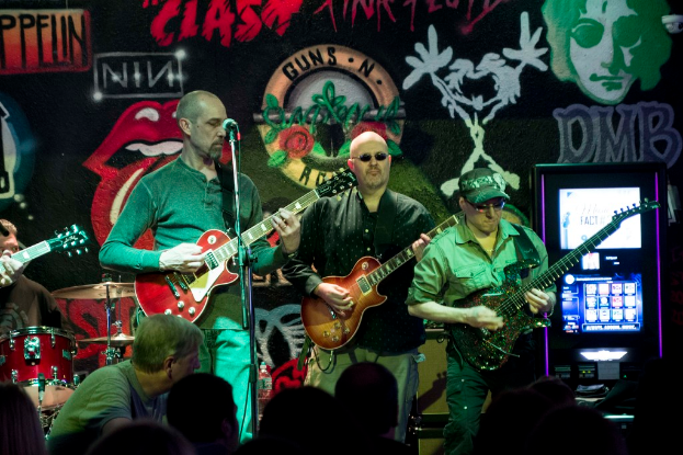 Drei Männer auf einer Bühne, die Gitarren spielen, mit einer Menge darunter und einer gemalten Wand im Hintergrund.