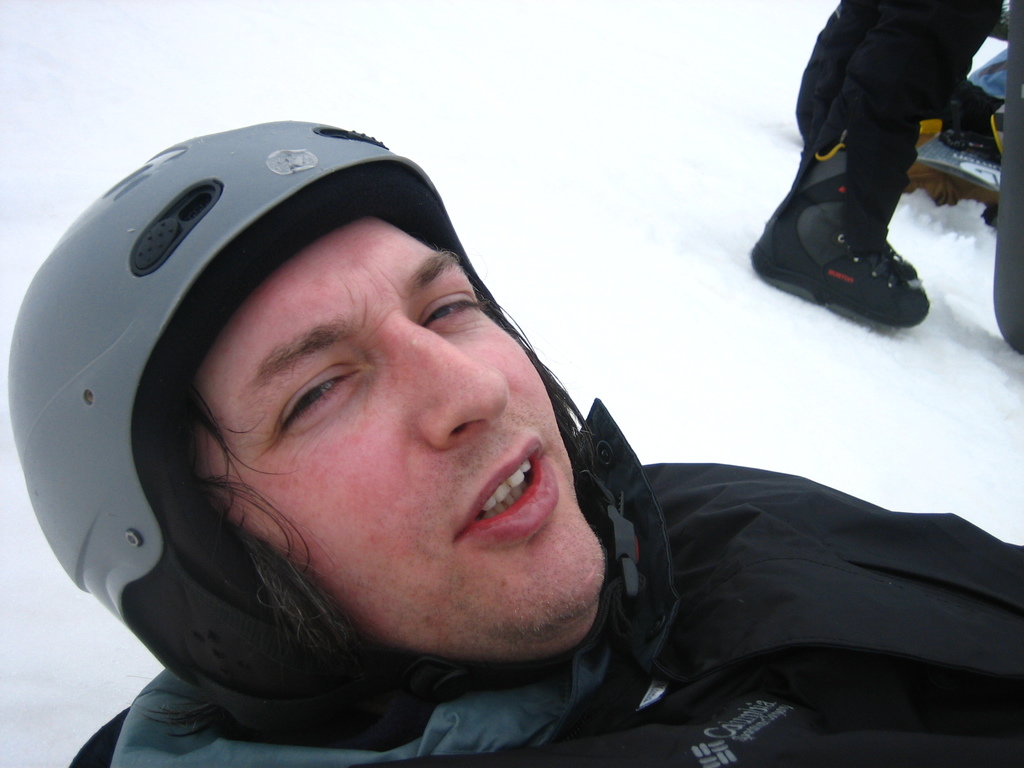 Ein Mann in Pullover und Helm steht im Schnee, mit den Beinen und Skiern einer anderen Person oben rechts im Bild.