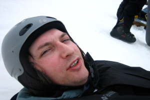 Ein Mann in Pullover und Helm steht im Schnee, mit den Beinen und Skiern einer anderen Person oben rechts im Bild.