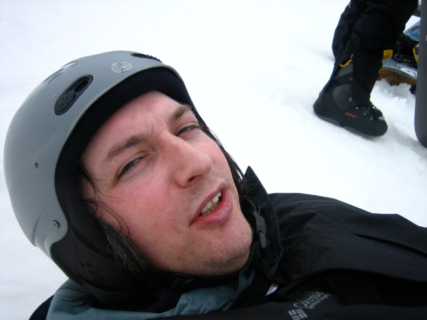 Ein Mann in Pullover und Helm steht im Schnee, mit den Beinen und Skiern einer anderen Person oben rechts im Bild.