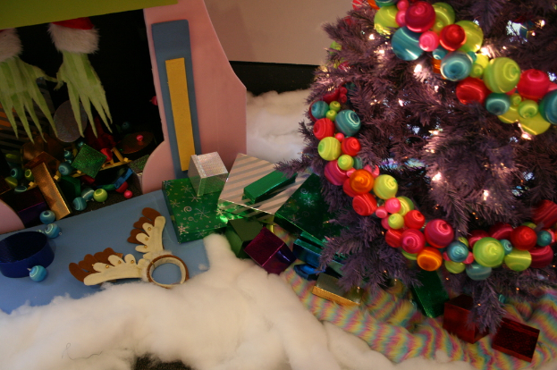 Drinnen steht ein geschmückter Weihnachtsbaum mit Lichterkette und Schmuck rechts, Geschenkboxen und Dekorationen in der Mitte, eine Hütte mit Dekorationen oben links und Baumwolle und Wolle unten.