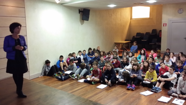 Eine Gruppe von Kindern, Jungen und Mädchen, sitzt auf dem Boden mit Büchern vor sich, während eine Frau links auf einer Bühne steht, mit einer Wand im Hintergrund.