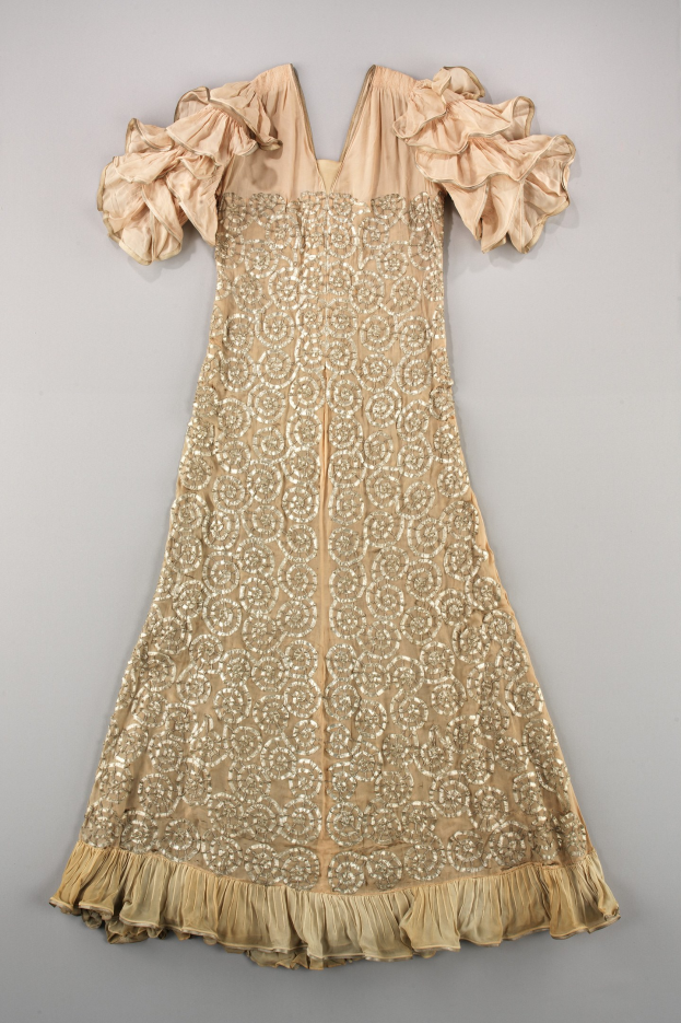 Ein cremefarbenes Kleid mit goldener Stickerei und Rüschenärmeln, das auf einer weißen Oberfläche mit detaillierten Designs und Mustern verziert ist.