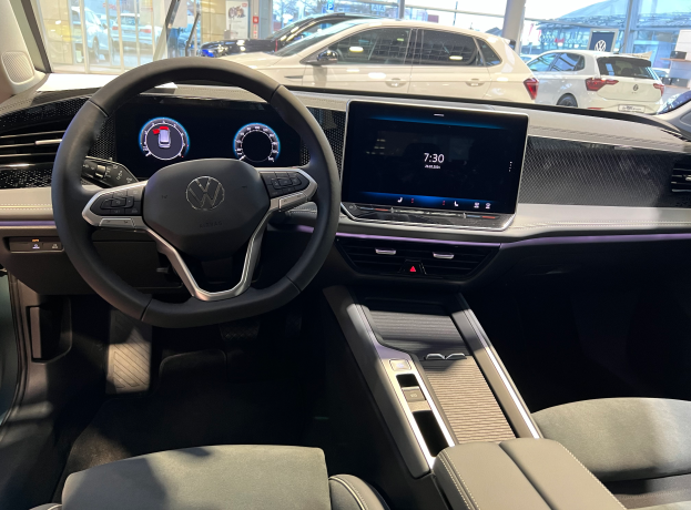 Das Innere eines Volkswagen e-Golf, mit Lenkrad, Armaturenbrett, Bildschirmen, Knöpfen, Sitzen und Glastüren, mit Blick auf andere Autos, Gebäude, Bäume, Pfosten und den Himmel durch die Glastüren.