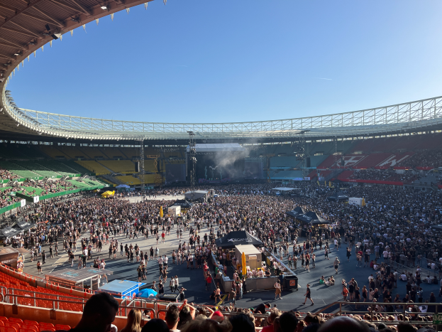 Eine große Menschenmenge versammelte sich im Wembley-Stadion für ein Konzert, mit einer Bühne, die verschiedene Gegenstände im Zentrum und ein Dach mit Lichtern an der Spitze des Bildes zeigt.