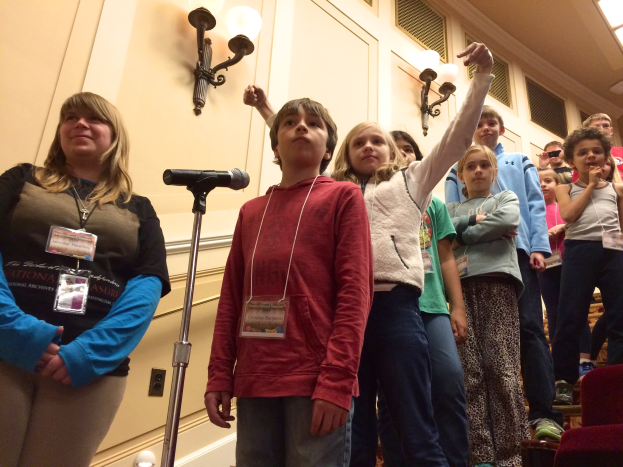 Gruppe von Kindern, die vor einem Mikrofon singen, mit einer Frau neben ihnen, Stühlen auf der rechten Seite und beleuchteter Wand im Hintergrund.