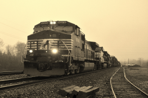 Ein Zug auf Bahnschienen mit Autos, Strommasten, Bäumen und einem Himmel im Hintergrund.
