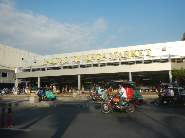 Eine belebte Straßenszene am Pasig City Mega Market mit Fahrzeugen und Fußgängern, ein Gebäude mit Text im Hintergrund und eine bewölkte Himmel.