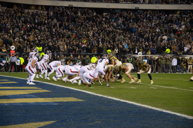 Ein Fußballspiel in einem Stadion mit Spielern in Sportkleidung und Helmen auf dem Feld, Zuschauern, Geländern und Tafeln mit Text im Hintergrund.