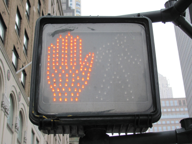 Rote Hand am Verkehrssignal in einer städtischen Umgebung mit Gebäuden auf beiden Seiten und einem klaren Himmel.