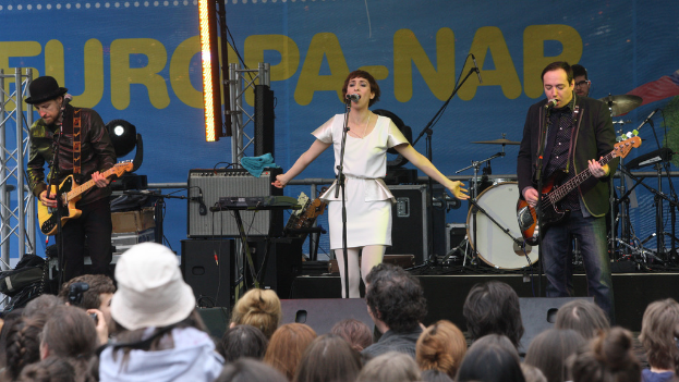 Eine Frau in einem weißen Kleid singt auf der Bühne bei einem Konzert, flankiert von zwei Gitarristen, mit einer Menge vorne und Lautsprechern im Hintergrund.