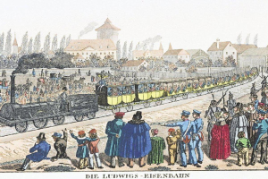 Eine Gruppe von Menschen, die um und in einem Zug auf Schienen stehen, mit Gebäuden und Bäumen im Hintergrund und Text unten, der "Ludwigs-Eisenbahn, der erste Bahnhof in Deutschland" liest.
