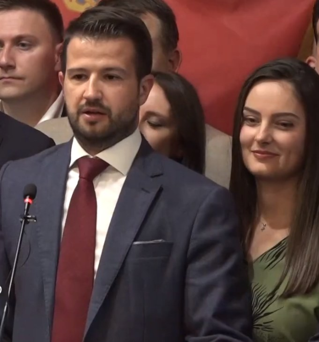 Ein Mann im Anzug spricht in ein Mikrofon an einem Rednerpult, mit einem Banner im Hintergrund und einer sichtbaren Menge, wahrscheinlich spricht er über ein Regierungsverbot der sozialen Medien.