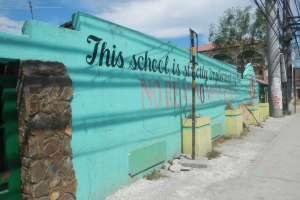 Eine grüne Wand mit Graffiti, das 'Diese Schule ist keine Stadt' lautet, umgeben von Gebäuden, Bäumen, Pfählen, Drähten, Brettern und anderen Gegenständen unter einem bewölkten Himmel.