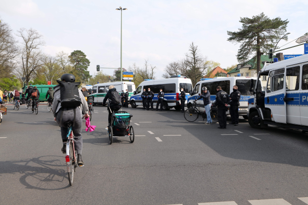 Eine Gruppe von Menschen, die auf Fahrrädern eine Straße entlangfahren, mit Polizeifahrzeugen, anderen Fahrzeugen, Bäumen und Gebäuden im Hintergrund.