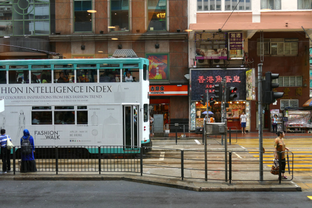 Ein Doppeldeckerbus fährt auf einer Straße mit hohen Gebäuden entlang, mit Fußgängern auf dem Gehweg, einem Geländer, Verkehrsampeln und Schildern mit Text und Bildern im Hintergrund.