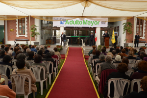 Ein Mann steht vor einem Gebäude mit einem Bogen, auf dem "Adulto Mayor" steht, während viele Menschen im Hintergrund auf Stühlen sitzen.