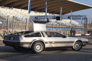 Ein silberner DeLorean DMC-12 Wagen befindet sich auf einer Straße umgeben von Menschen, Flaggen, Schildern, Geländern, Stühlen und einem Schuppen, mit Bäumen und einem klaren blauen Himmel im Hintergrund.