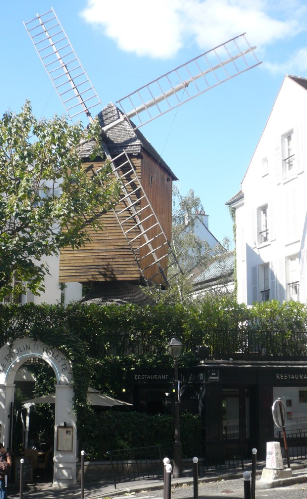Eine Windmühle, mehrere Gebäude, Bäume, Metallstangen und ein Zaun in ländlicher Umgebung.