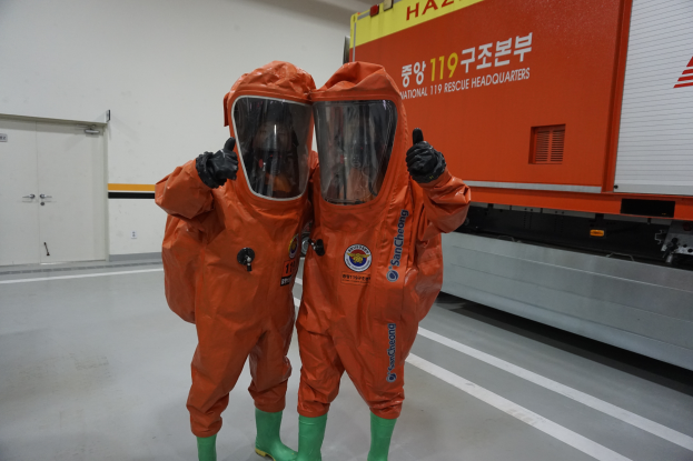 Zwei Personen in orangen Chemikalienschutzanzügen stehen nebeneinander vor einem Lastwagen, mit einer Wand und Türen im Hintergrund, was darauf hinweist, dass sie Teil des Nationalen Einsatzteams sind.