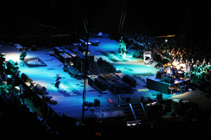 Eine Gruppe von Menschen, die auf einer Bühne mit Lautsprechern, Scheinwerfern und Mikrofonen performen, umgeben von einer großen Menge in einem dunklen Hintergrund.