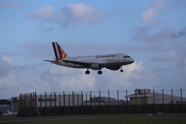 Germanwings Airbus A320-200 beim Starten am Frankfurter Flughafen mit Gras darunter, einem Zaun im Vordergrund, Gebäuden und Bäumen im Hintergrund und einer bewölkten Himmel
