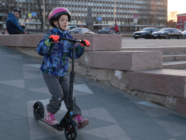 Ein junger Junge fährt mit einem Helm und Handschuhen auf einem Scooter eine Straße entlang, mit verschiedenen städtischen Elementen und einem klaren blauen Himmel im Hintergrund.