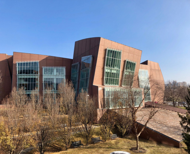 Großes Gebäude mit zahlreichen Fenstern, umgeben von Bäumen, Bänken, Gras und einem Laternenmast, mit anderen Gebäuden und einem klaren blauen Himmel im Hintergrund, als der neue Campus der University of Illinois at Chicago identifiziert.