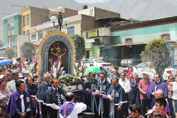 Menschen auf einer Straße vor der Stadt, mit einem Kruzifix und Blumensträußen im Vordergrund, fahrende Autos im Hintergrund und Gebäude, Bäume und Straßenlaternen weiter hinten.
