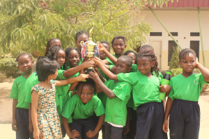 Eine Gruppe von Kindern hält einen Pokal in einem Schulhof mit Bäumen, Pflanzen und einem Gebäude im Hintergrund.