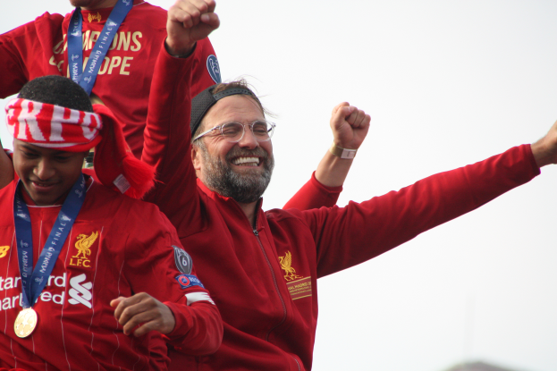 Liverpool-Trainer Jürgen Klopp feiert mit seinen Spielern nach dem Gewinn der UEFA Champions League, während er eine rote Jacke trägt und von zwei medalien tragenden Personen umgeben ist. Der Himmel ist im Hintergrund zu sehen.
