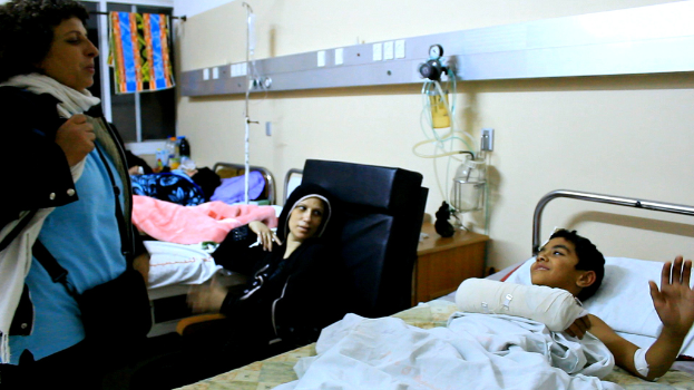 Ein Krankenhauszimmer mit mehreren Betten, auf denen jeweils ein Patient liegt, und eine Frau, die in der Nähe eines der Betten steht und eine Tasche trägt.