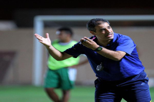 Ein Mann in einem blauen T-Shirt und blauen Hosen steht auf einem Fußballfeld und hält ein Mikrofon in der Hand, während er seinen Spielern Zeichen gibt. Im Hintergrund ist eine weitere Person in einem grünen Kleid, ein Torpfosten und eine Wand zu sehen.
