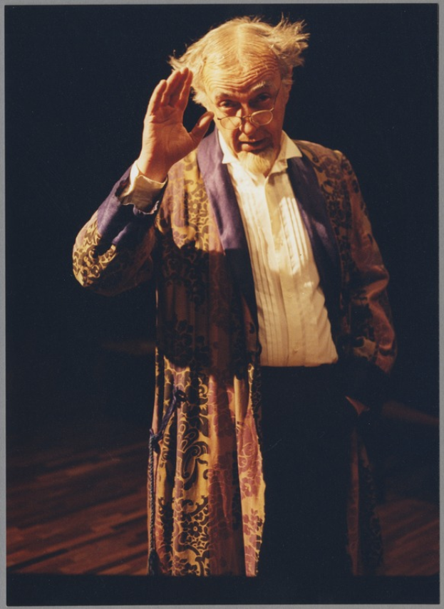 Ein Mann in einem Gewand und Brille steht auf einer Bühne mit dunklem Hintergrund und schaut ernst in die Menge.