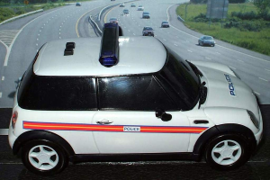 Ein weißer Polizeiwagen steht im Vordergrund mit vielen Autos auf der Straße dahinter, flankiert von Gras und Bäumen auf beiden Seiten in einer ländlichen Umgebung.